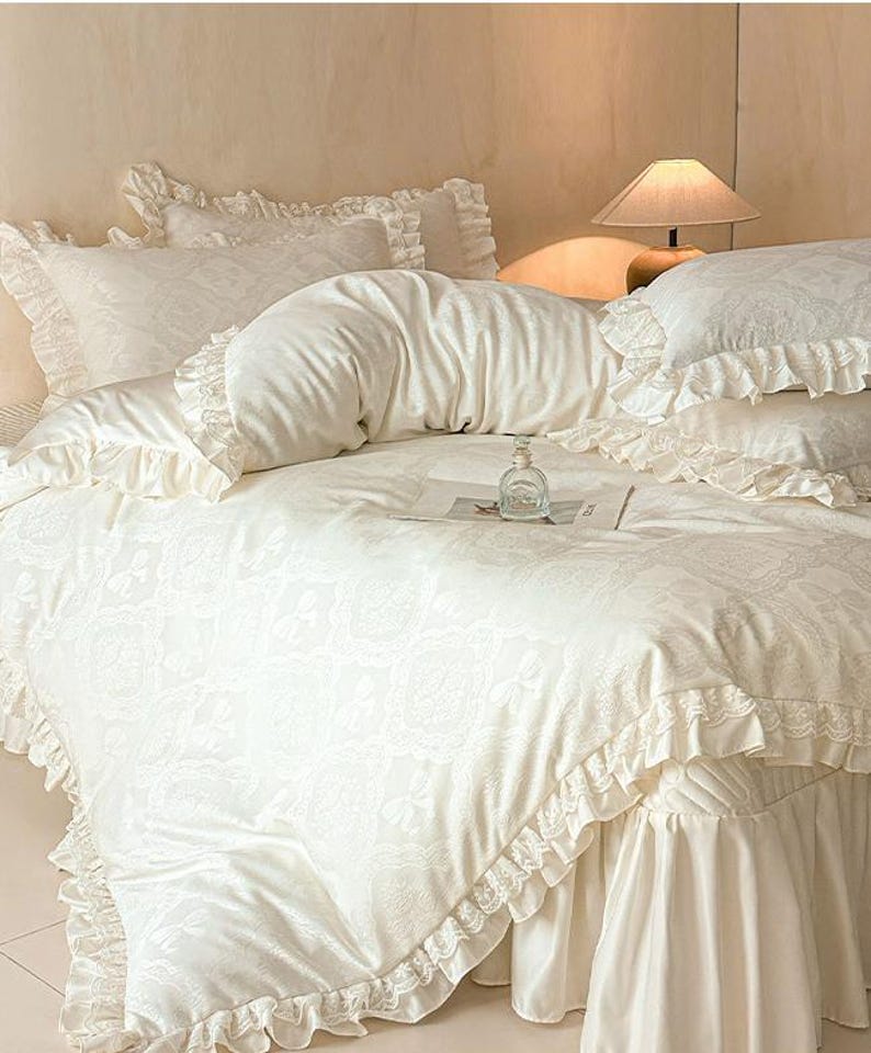 K&ouml;nnte beinhalten: Ein cremefarbenes Bettw&auml;sche-Set bestehend aus einem Bettbezug, passenden Kissenbez&uuml;gen und einem Bettvolant, verziert mit einem dezenten, tonalen floralen Jacquardmuster und zarten R&uuml;schenkanten. Eine kleine, klare Glasflasche steht auf dem Bett.