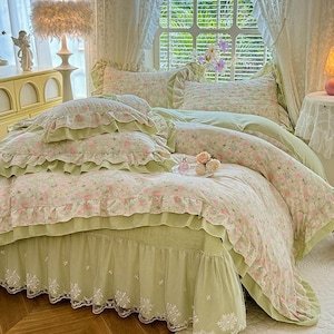 Può includere: Un set di biancheria da letto a fantasia floreale nei toni del verde e del rosa. Il set comprende un piumino, cuscini e una gonna da letto, tutti adornati da bordi a balze. La biancheria da letto è disposta su un letto, creando un'atmosfera accogliente.