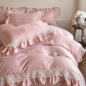 Puede incluir: Un juego de cama rosa claro con un sutil patr&oacute;n jacquard de medallones florales y lazos. La funda n&oacute;rdica y las fundas de almohada est&aacute;n adornadas con ribetes de encaje blanco y bordes con volantes. Un peque&ntilde;o frasco de vidrio descansa sobre la tela.