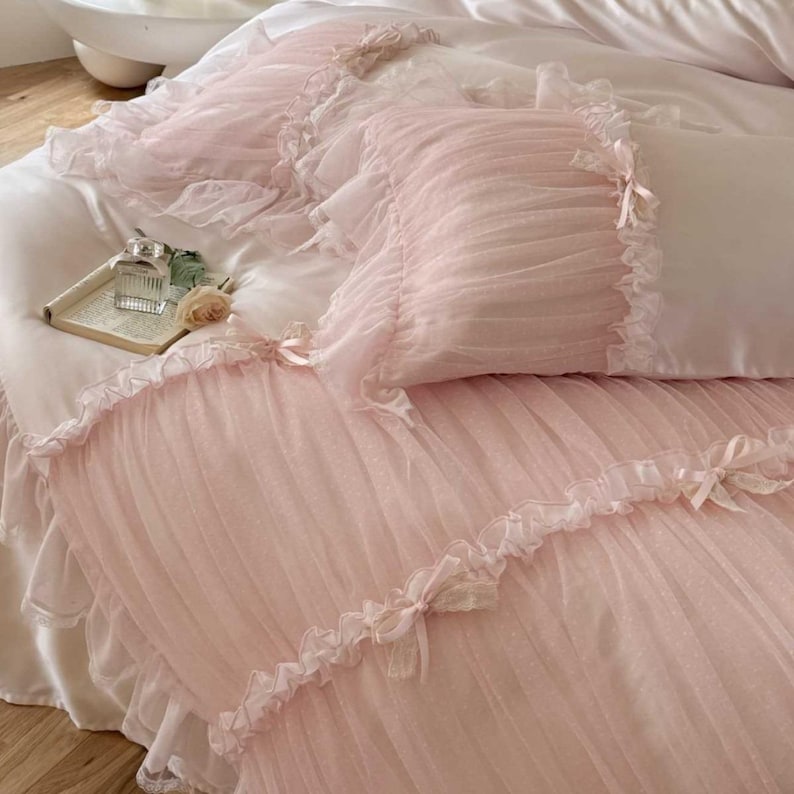 May include: A light pink bed set with a ruffled, sheer overlay. The set includes a comforter and pillowcases, all adorned with delicate bows. The bedding is arranged on a white bed, creating a soft and romantic aesthetic.