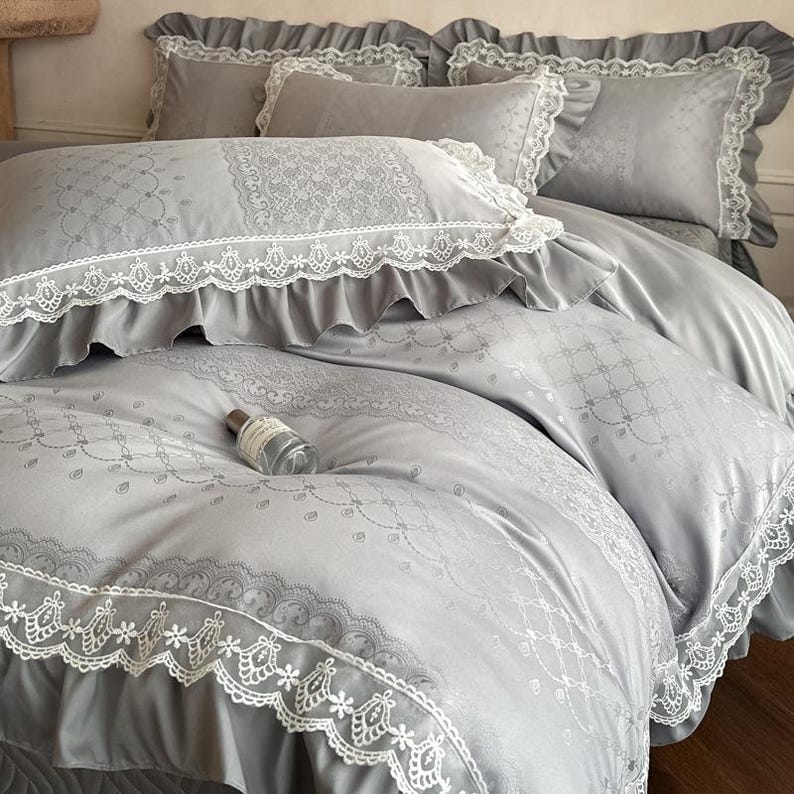 Puede incluir: Un juego de cama gris claro que incluye una funda n&oacute;rdica y fundas de almohada a juego con ribetes de encaje blanco y volantes. La tela presenta un sutil patr&oacute;n de damasco tonal con intrincados motivos florales y de celos&iacute;a en la superficie.