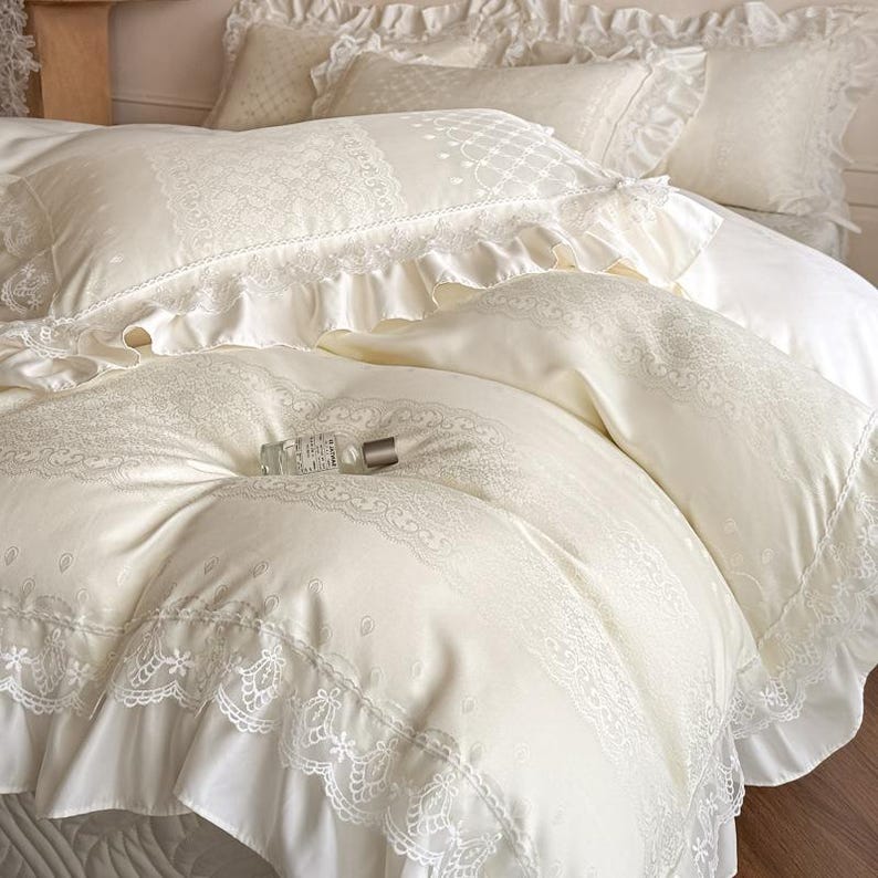 Puede incluir: Un juego de cama de color crema que incluye funda n&oacute;rdica y fundas de almohada con ribetes de encaje, bordes con volantes y un sutil patr&oacute;n floral. Un peque&ntilde;o frasco de vidrio transparente con tapa plateada descansa sobre la funda n&oacute;rdica.