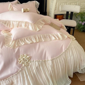 May include: A light pink bedding set featuring a duvet cover and pillowcases with cream-colored ruffled edges. The duvet cover is adorned with large, three-dimensional fabric flower appliqués in a matching cream tone, adding texture to the smooth surface.