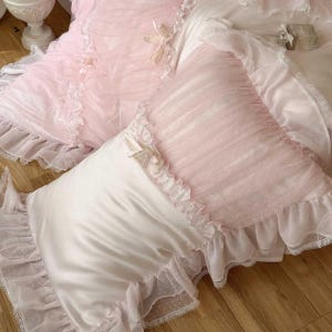 May include: A close-up of a bed with a white satin pillow and a pink pillow with a ruffled edge. The pink pillow has a sheer overlay with white polka dots. The bedding has a romantic, feminine aesthetic.