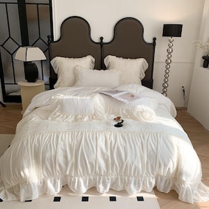 Puede incluir: Un juego de cama color crema con una funda nórdica con volantes y fundas de almohada a juego. La ropa de cama presenta un estampado floral texturizado y está colocada sobre una cama con un cabecero marrón oscuro. Se ve una lámpara negra y una lámpara de pie.