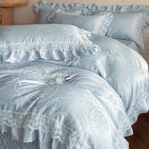 Puede incluir: Un juego de cama azul claro con acabado satinado y un patr&oacute;n repetitivo de medallones florales y lazos. La funda n&oacute;rdica y las fundas de almohada est&aacute;n decoradas con bordes de encaje blanco y volantes, creando una apariencia texturizada.