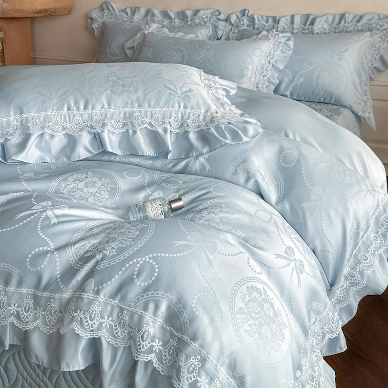 Puede incluir: Un juego de cama azul claro con acabado satinado y un patr&oacute;n repetitivo de medallones florales y lazos. La funda n&oacute;rdica y las fundas de almohada est&aacute;n decoradas con bordes de encaje blanco y volantes, creando una apariencia texturizada.