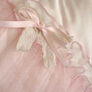 May include: Close-up of a delicate, light pink fabric with a white lace trim and a matching pink ribbon tied in a bow. The fabric has a dotted pattern, and the overall aesthetic is soft and feminine.
