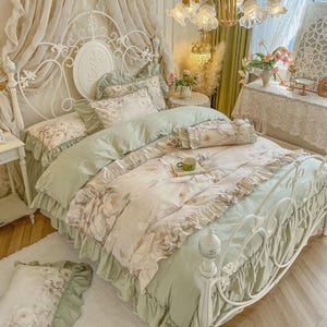 Peut inclure: Un lit en métal blanc avec une literie florale dans des tons vert clair et crème. La housse de couette et les oreillers présentent un motif floral et des bords à volants. Une petite tasse verte repose sur un plateau sur le lit.