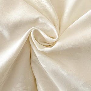 K&ouml;nnte beinhalten: Ein cremefarbener Stoff mit einem dezenten, Ton-in-Ton gehaltenen floralen Damastmuster. Das Material ist in der Mitte gerafft, wodurch weiche, flie&szlig;ende Falten und Lichtreflexe entstehen, die die glatte, leicht gl&auml;nzende Textur der Oberfl&auml;che betonen.