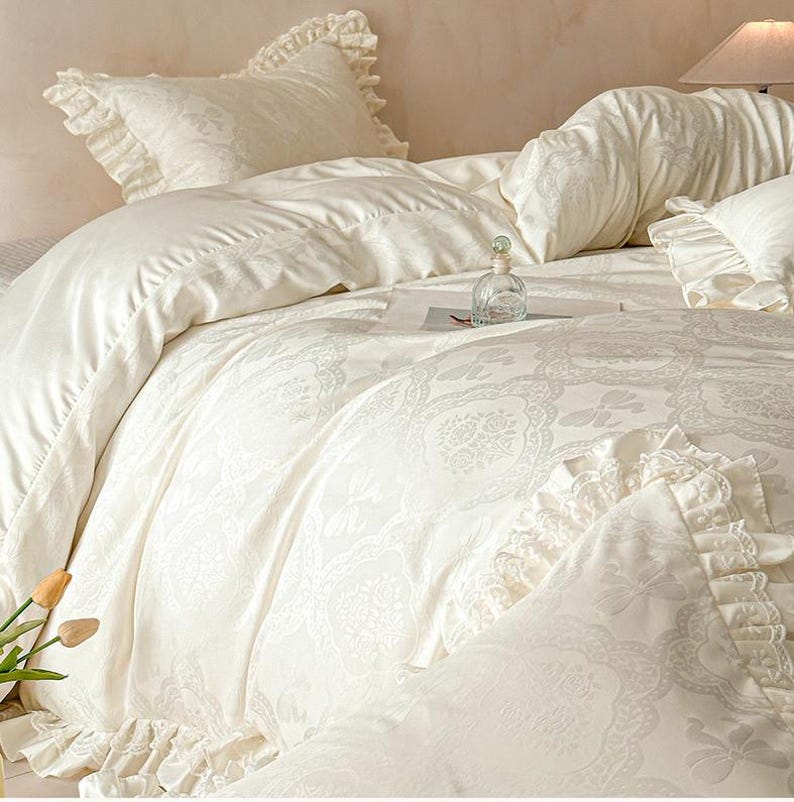 K&ouml;nnte beinhalten: Ein cremefarbenes Bettw&auml;sche-Set bestehend aus einem Bettbezug und Kissenbez&uuml;gen mit einem tonalen Damastmuster und ger&uuml;schten Kanten. Der Stoff zeigt ein dezentes Jacquard-Gewebe mit floralen Motiven und Schmetterlingsmustern.