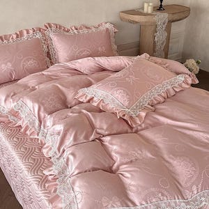 Puede incluir: Un juego de cama rosa claro con acabado satinado y un patr&oacute;n repetitivo de lazos y medallones florales. La funda n&oacute;rdica y las fundas de almohada est&aacute;n adornadas con encaje blanco y bordes con volantes. El fald&oacute;n tiene una textura ondulada.