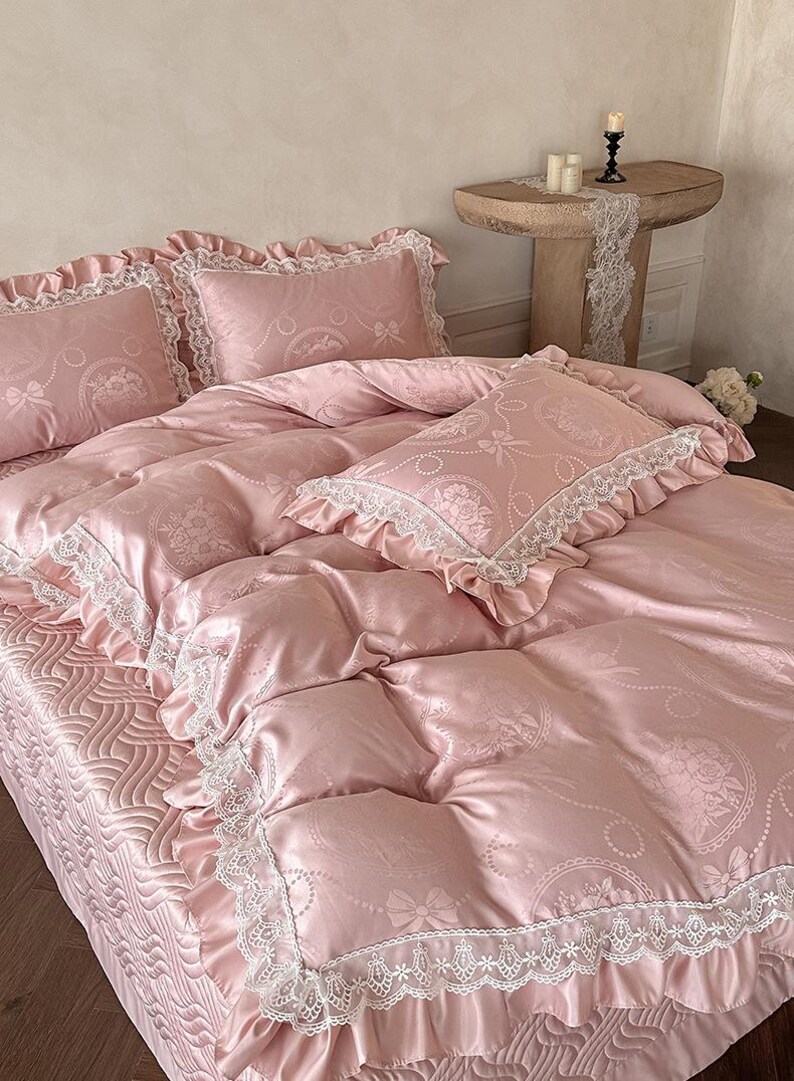 Puede incluir: Un juego de cama rosa claro con acabado satinado y un patr&oacute;n repetitivo de lazos y medallones florales. La funda n&oacute;rdica y las fundas de almohada est&aacute;n adornadas con encaje blanco y bordes con volantes. El fald&oacute;n tiene una textura ondulada.