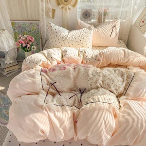 May include: A cozy bed with a light pink comforter and pillows. The comforter features floral and lace details, with a soft, textured center. Pillows are patterned with small black shapes. The scene is bathed in soft, natural light, creating a warm and inviting atmosphere.