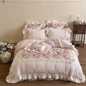 May include: A light pink floral bedding set with ruffled edges. The set includes a comforter, pillows, and decorative cushions. The comforter features a floral pattern with lace trim. The pillows and cushions have ruffled edges and floral accents.