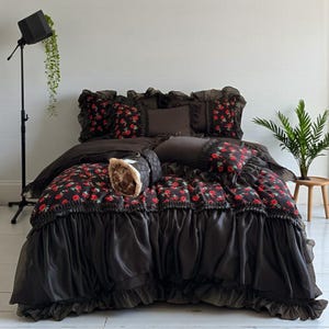 May include: A dark grey bed linen set featuring a floral pattern of red roses. The set includes a duvet cover, pillows, and a decorative bolster cushion. The duvet cover has a ruffled edge, and the pillows have a matching floral design.
