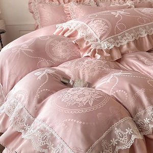 Puede incluir: Un juego de cama rosa suave con un patr&oacute;n repetitivo de medallones florales, lazos y l&iacute;neas punteadas. La funda n&oacute;rdica y las fundas de almohada est&aacute;n rematadas con un borde de encaje blanco ancho e intrincado. Una peque&ntilde;a botella de vidrio descansa encima.