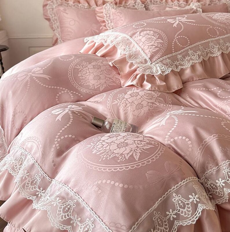 Puede incluir: Un juego de cama rosa suave con un patr&oacute;n repetitivo de medallones florales, lazos y l&iacute;neas punteadas. La funda n&oacute;rdica y las fundas de almohada est&aacute;n rematadas con un borde de encaje blanco ancho e intrincado. Una peque&ntilde;a botella de vidrio descansa encima.