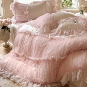 May include: A pink bedding set with a romantic, feminine aesthetic. The set includes a comforter, pillows, and a bed skirt, all adorned with ruffles and delicate bows. The comforter has a sheer, layered design, creating a soft, airy look.