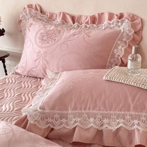 Puede incluir: Un juego de cama rosa empolvado que incluye dos fundas de almohada con ribete de encaje blanco y bordes con volantes. La tela tiene un sutil patr&oacute;n jacquard de medallones florales y lazos. Una botella de vidrio transparente sobre la cama.