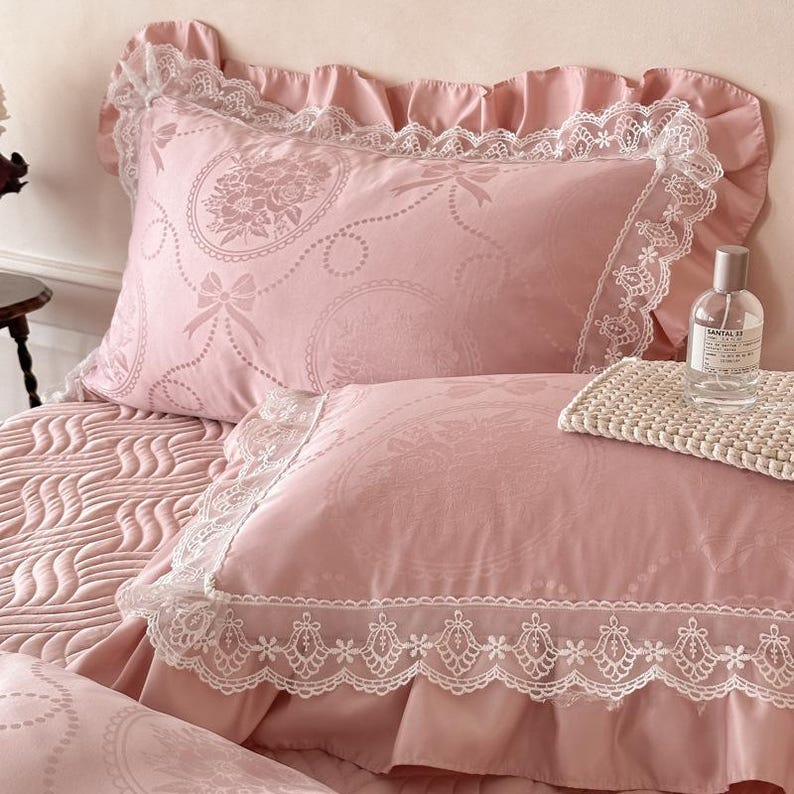 Puede incluir: Un juego de cama rosa empolvado que incluye dos fundas de almohada con ribete de encaje blanco y bordes con volantes. La tela tiene un sutil patr&oacute;n jacquard de medallones florales y lazos. Una botella de vidrio transparente sobre la cama.