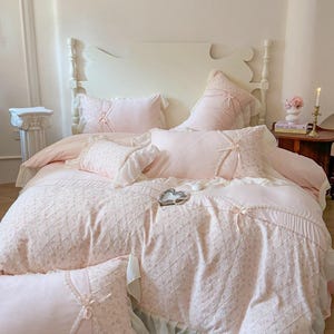 May include: A light pink bed set with a white headboard. The bedding includes a comforter, pillows, and shams, all in a soft pink hue with delicate lace and ribbon details. The comforter features a diamond pattern.
