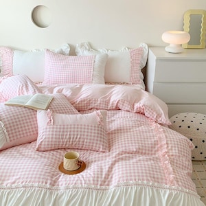 May include: A bedding set featuring a light pink and white gingham pattern with white ruffled trim. The set includes a duvet cover, two standard pillowcases, and a rectangular accent pillow, all decorated with delicate ruffles along the edges.