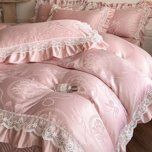 Puede incluir: Un juego de cama rosa claro con acabado satinado y un patr&oacute;n repetitivo de rosas en marcos circulares y lazos. La funda n&oacute;rdica y las fundas de almohada est&aacute;n adornadas con bordes de encaje blanco y volantes. Una botella de vidrio sobre la cama.