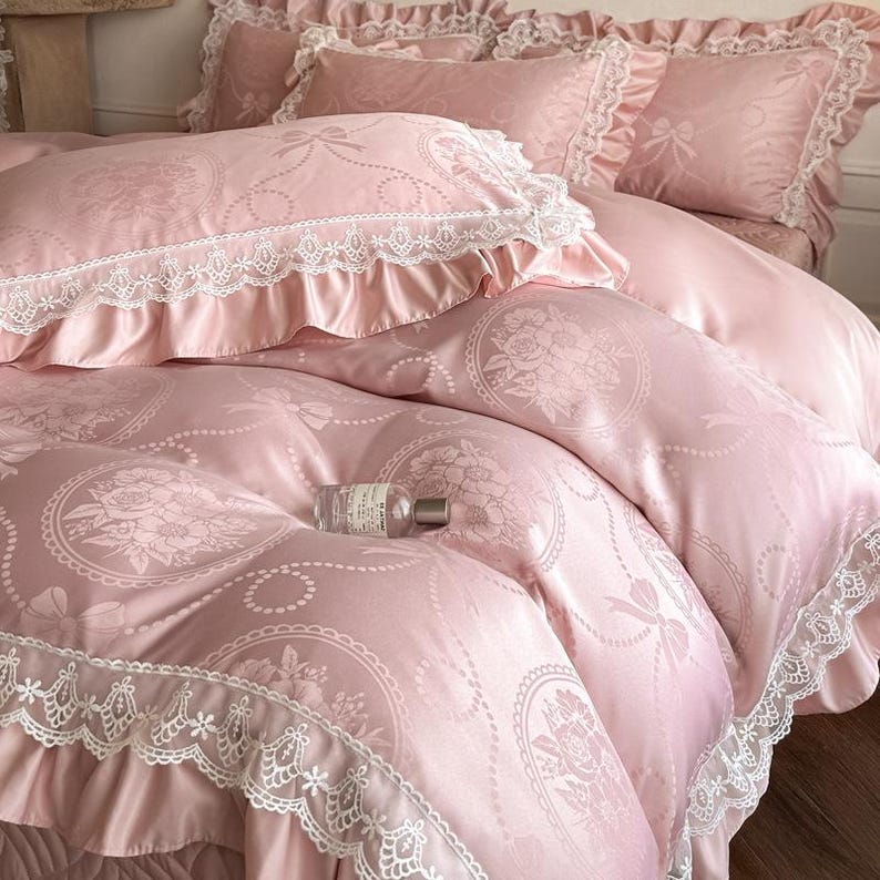 Puede incluir: Un juego de cama rosa claro con acabado satinado y un patr&oacute;n repetitivo de rosas en marcos circulares y lazos. La funda n&oacute;rdica y las fundas de almohada est&aacute;n adornadas con bordes de encaje blanco y volantes. Una botella de vidrio sobre la cama.