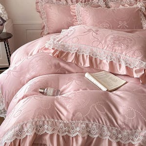 Puede incluir: Un juego de cama rosa claro con un sutil patr&oacute;n jacquard de lazos y medallones florales. La funda n&oacute;rdica y las fundas de almohada tienen volantes de encaje blanco. Un libro abierto y una peque&ntilde;a botella de vidrio descansan sobre la tela.