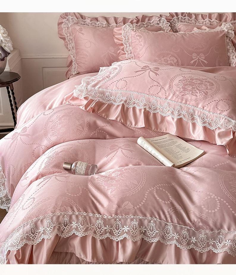 Puede incluir: Un juego de cama rosa claro con un sutil patr&oacute;n jacquard de lazos y medallones florales. La funda n&oacute;rdica y las fundas de almohada tienen volantes de encaje blanco. Un libro abierto y una peque&ntilde;a botella de vidrio descansan sobre la tela.