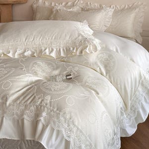 Puede incluir: Un juego de cama de color crema que incluye funda n&oacute;rdica y fundas de almohada con un patr&oacute;n tonal de flores y lazos. Los bordes est&aacute;n rematados con encaje con volantes. Un peque&ntilde;o frasco de vidrio con tapa plateada descansa sobre el edred&oacute;n.