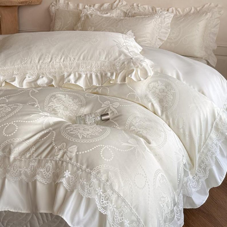 Puede incluir: Un juego de cama de color crema que incluye funda n&oacute;rdica y fundas de almohada con un patr&oacute;n tonal de flores y lazos. Los bordes est&aacute;n rematados con encaje con volantes. Un peque&ntilde;o frasco de vidrio con tapa plateada descansa sobre el edred&oacute;n.
