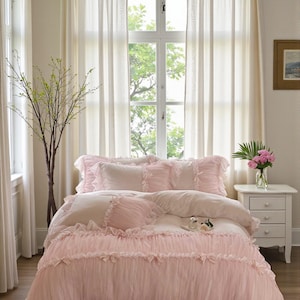 May include: A bed with a pink ruffled comforter and matching pillows. The bedding has a delicate, feminine aesthetic. The room features sheer white curtains, a small white nightstand, and a vase of pink flowers, creating a soft, inviting atmosphere.