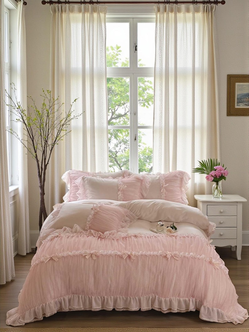 May include: A bed with a pink ruffled comforter and matching pillows. The bedding has a delicate, feminine aesthetic. The room features sheer white curtains, a small white nightstand, and a vase of pink flowers, creating a soft, inviting atmosphere.