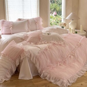 May include: A bed with a white fitted sheet and a pink ruffled comforter and pillow set. The comforter and pillows have a delicate, sheer overlay with ruffled edges. The room has a window with natural light and a white lamp.