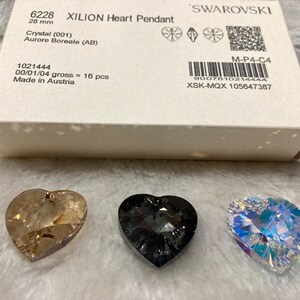 May include: Three crystal heart pendants in different colors: a light brown, a dark gray, and a clear iridescent. The pendants are on a white surface. The text on the box reads "6228 XILION Heart Pendant 28 mm Crystal (001) Aurore Boreale (AB) 1021444 00/01/04 gross = 16 pcs Made in Austria M-P4-C4 9007810214444 XSK-MQX 105647387 SWAROVSKI"