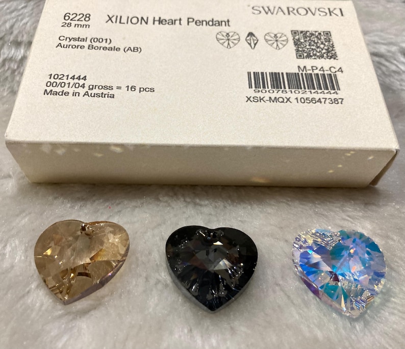 May include: Three crystal heart pendants in different colors: a light brown, a dark gray, and a clear iridescent. The pendants are on a white surface. The text on the box reads "6228 XILION Heart Pendant 28 mm Crystal (001) Aurore Boreale (AB) 1021444 00/01/04 gross = 16 pcs Made in Austria M-P4-C4 9007810214444 XSK-MQX 105647387 SWAROVSKI"