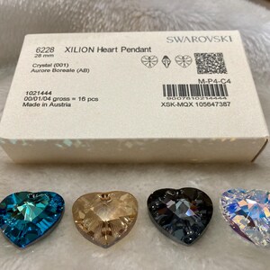 May include: Four heart-shaped crystal pendants in different colors: blue, brown, gray, and clear. The pendants are on a white surface. The box they came in is visible in the background. The box is white with black text that reads "6228 XILION Heart Pendant 28 mm Crystal (001) Aurore Boreale (AB) 1021444 00/01/04 gross = 16 pcs Made in Austria M-P4-C4 9007610214444 XSK-MQX 105647387 SWAROVSKI"