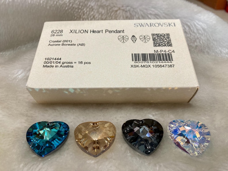 May include: Four heart-shaped crystal pendants in different colors: blue, brown, gray, and clear. The pendants are on a white surface. The box they came in is visible in the background. The box is white with black text that reads "6228 XILION Heart Pendant 28 mm Crystal (001) Aurore Boreale (AB) 1021444 00/01/04 gross = 16 pcs Made in Austria M-P4-C4 9007610214444 XSK-MQX 105647387 SWAROVSKI"