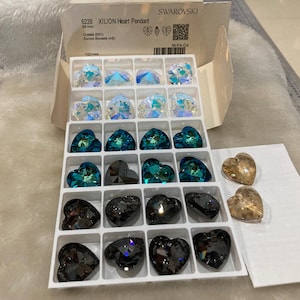 May include: A clear plastic box filled with various heart-shaped crystal pendants. The box is labeled "6228 XILION Heart Pendant 20 mm Crystal 2011 Autore Boreale (A)" and "SWAROVSKI". There are also two smaller boxes with additional heart-shaped pendants.