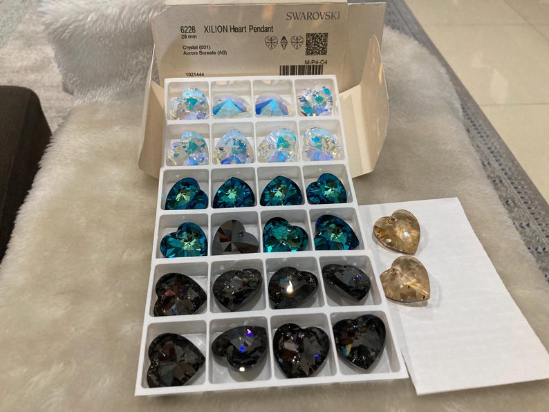 May include: A clear plastic box filled with various heart-shaped crystal pendants. The box is labeled "6228 XILION Heart Pendant 20 mm Crystal 2011 Autore Boreale (A)" and "SWAROVSKI". There are also two smaller boxes with additional heart-shaped pendants.