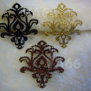 May include: Three decorative, glitter-covered, paper cutouts in the shape of a damask design. The cutouts are black, gold, and burgundy.