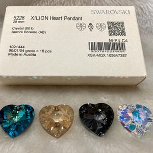May include: Four heart-shaped crystal pendants in different colors: blue, beige, gray, and iridescent. The pendants are on a white surface. The packaging for the pendants is in the background. The packaging is white with the text "Swarovski" and "Xilion Heart Pendant" printed on it.