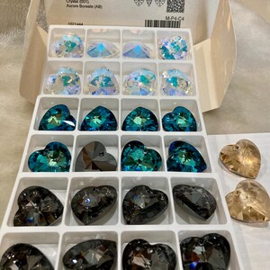May include: A white box with a clear plastic tray containing 18 heart-shaped crystal pendants. The box has the text "6228 XILION Heart Pendant 28 mm Crystal (001) Aurore Boreale (AB) 1001444 SWAROVSKI M-P4-C4" printed on it.
