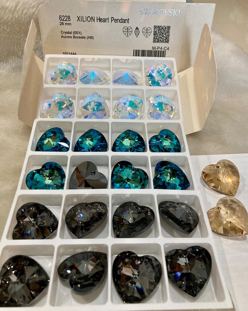 May include: A white box with a clear plastic tray containing 18 heart-shaped crystal pendants. The box has the text "6228 XILION Heart Pendant 28 mm Crystal (001) Aurore Boreale (AB) 1001444 SWAROVSKI M-P4-C4" printed on it.