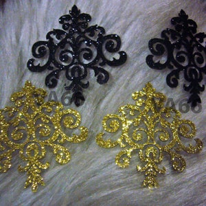 May include: Four decorative embellishments, two are gold glitter and two are black glitter. The embellishments are in the shape of a stylized floral design.