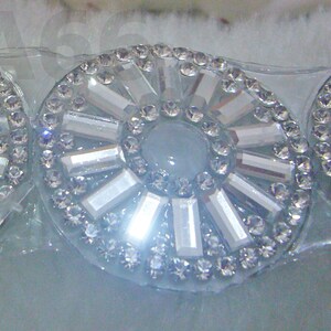 May include: A close-up of a decorative embellishment with a circular design. The embellishment is made of clear rhinestones and silver-toned rectangular pieces arranged in a radial pattern.