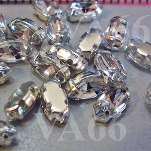 May include: A close-up of a pile of silver-toned, oval-shaped, crystal rhinestones with a hole in the center. The rhinestones are scattered on a light gray surface.