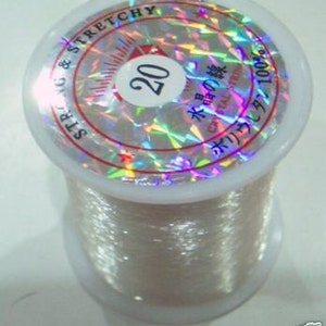 May include: A spool of clear, iridescent, stretchy string. The label reads "20 水晶の糸 Crystal String 米国より輸入 100% ポリエステル"